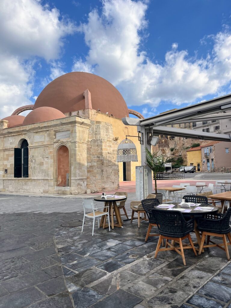 An outdoor café with modern black and wooden chairs sits on a stone-paved square, facing a historic domed building made of beige and terracotta-colored stone. The domes have a smooth, reddish-brown finish, contrasting with the textured stone walls. The sky is bright blue with scattered fluffy white clouds, and a few pigeons rest atop the structure. In the background, a mix of historic and modern buildings, as well as parked vehicles, add depth to the scene.