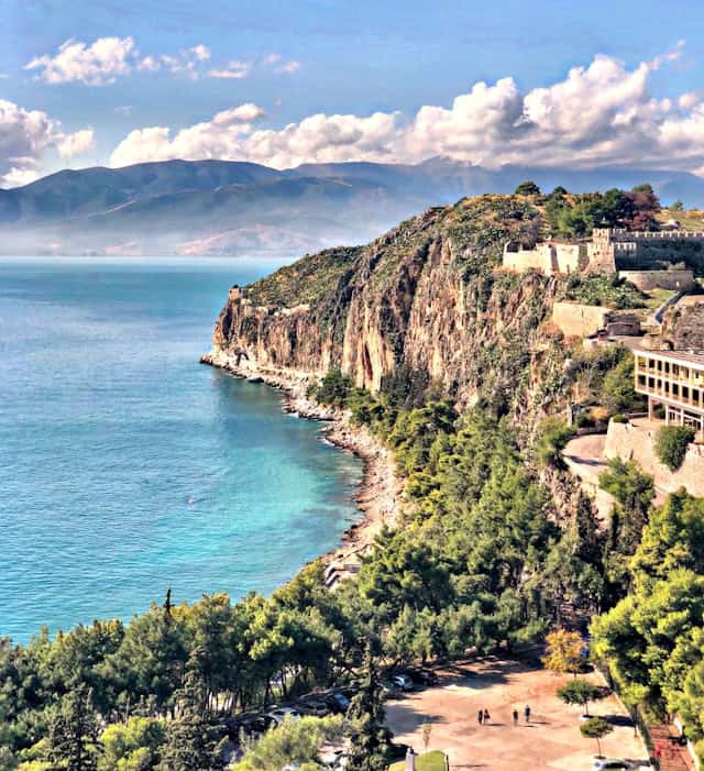 A breathtaking coastal view with turquoise waters meeting rugged cliffs covered in lush greenery. Atop the cliffs, a historic fortress stands, overlooking the serene sea. In the background, misty mountains stretch beneath a sky dotted with fluffy white clouds.