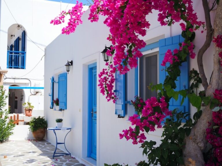 A charming Greek island street with white-washed houses featuring bright blue doors and shutters. Pink bougainvillea cascades over the walls, adding vibrant contrast to the crisp white architecture. The cobblestone path is adorned with small potted plants and a simple blue table.