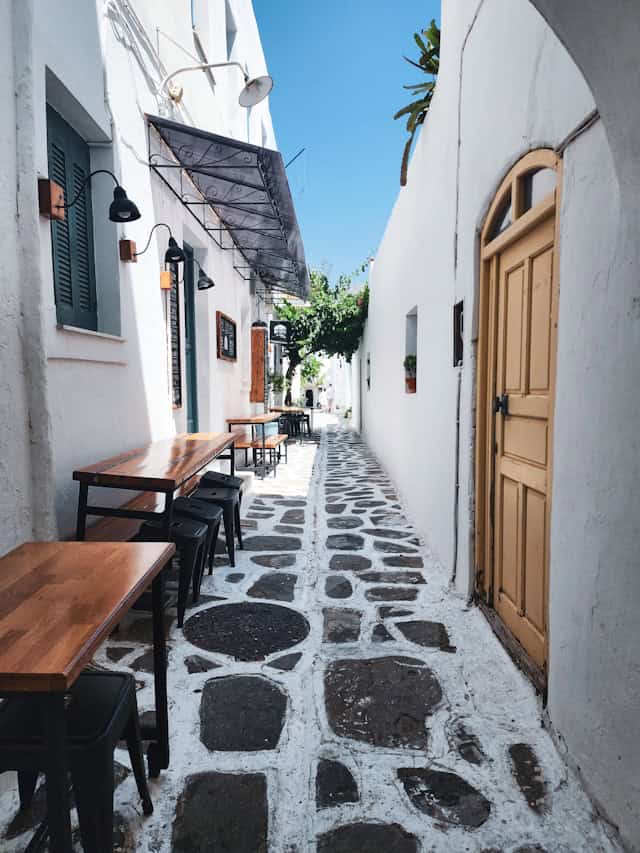 A charming narrow alleyway in a Mediterranean town, lined with white-washed buildings and rustic wooden doors. Outdoor café tables with black stools are set along the cobblestone pathway, which features an intricate stone and white grout pattern. A black wrought-iron awning and wall-mounted lamps add to the cozy ambiance, while lush greenery peeks through at the far end of the alley.