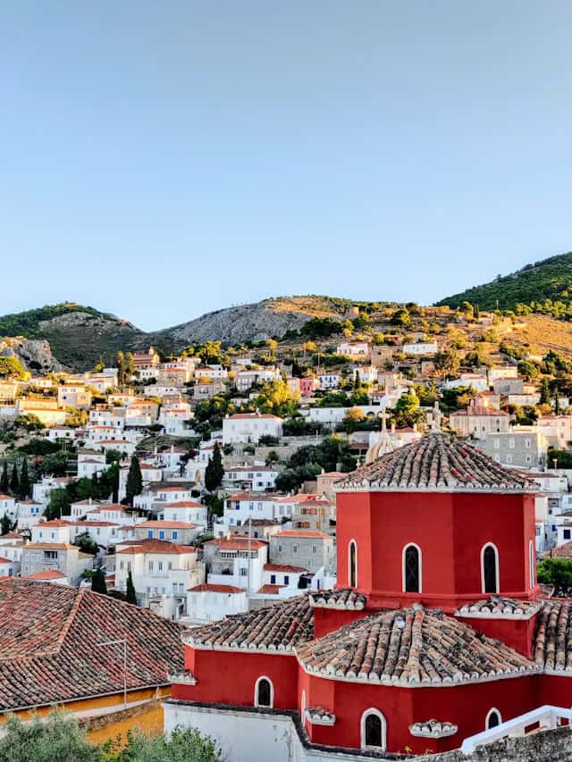 A picturesque hillside town with traditional Mediterranean architecture, where white and pastel-colored houses are nestled among lush greenery. In the foreground, a striking red-domed church with terracotta roof tiles stands out against the blue sky. Rolling hills and rugged mountains frame the idyllic landscape.