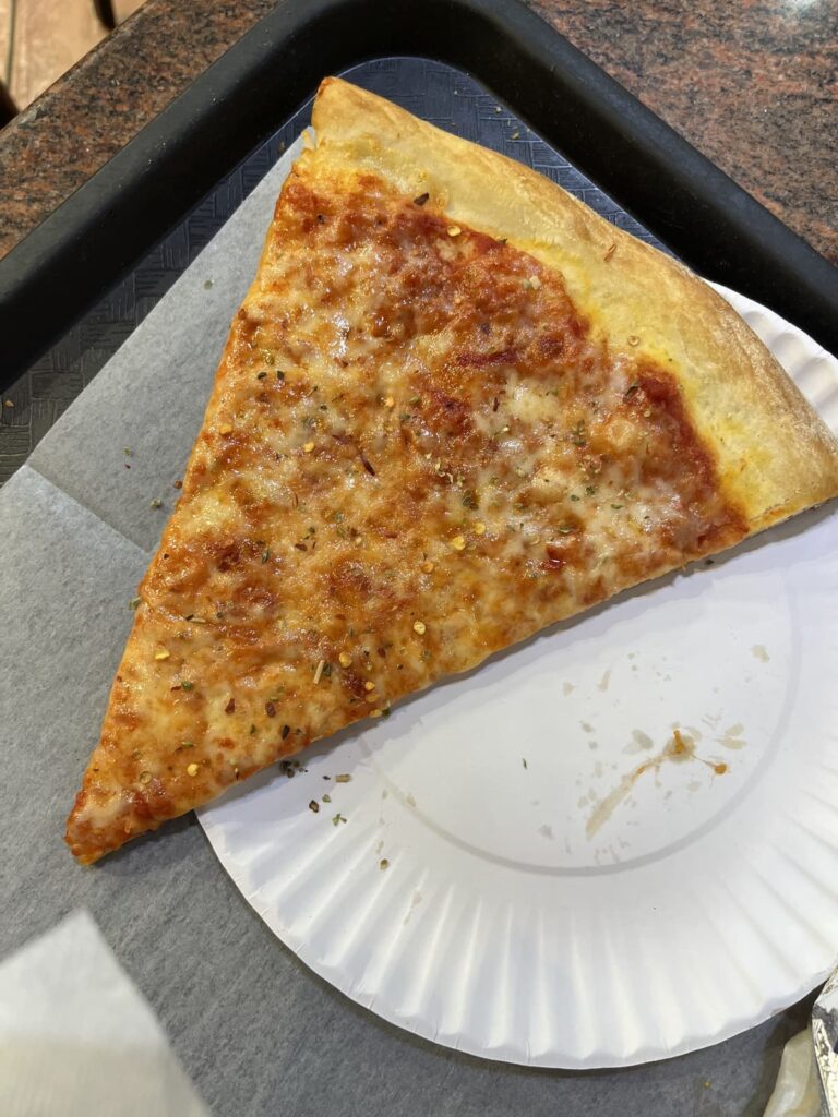 A slice of New York-style pizza on a white paper plate, placed on a black tray with a textured gray liner. The pizza is thin-crusted, topped with golden melted cheese and sprinkles of oregano and crushed red pepper flakes.