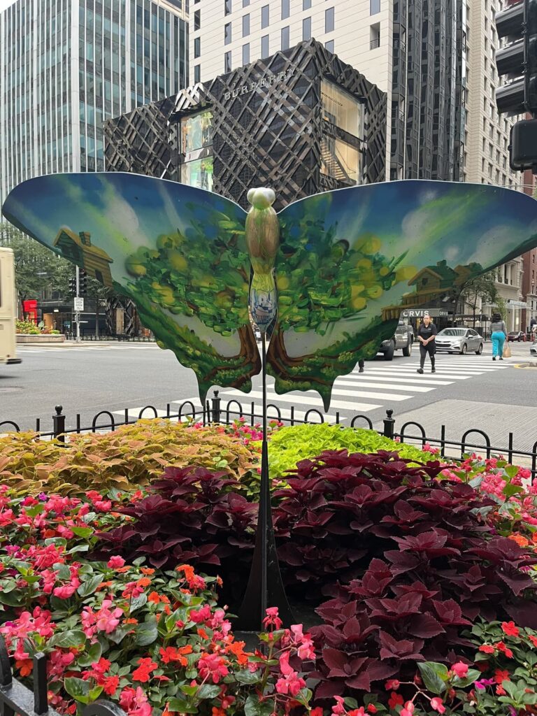 A colorful, oversized butterfly sculpture with painted nature scenes displayed among vibrant red, pink, and green flowers. The background shows a Burberry store with a geometric facade and modern skyscrapers in a bustling city.