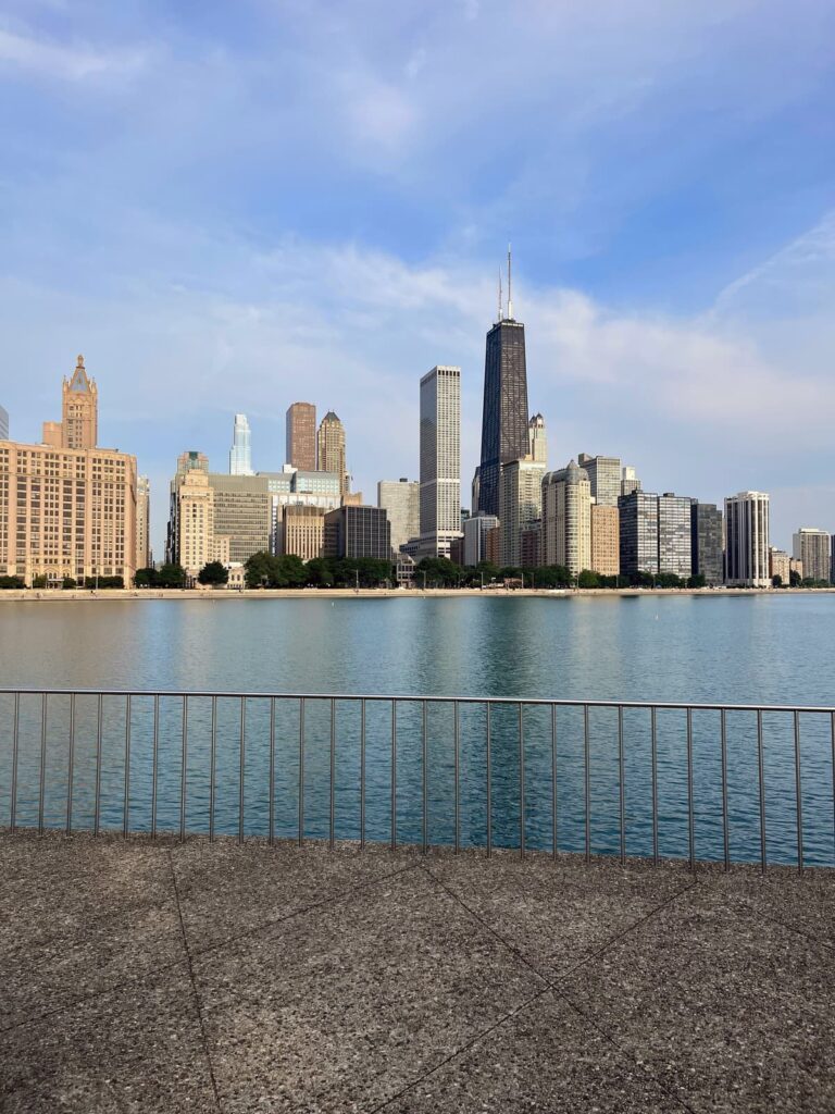 A waterfront view of Chicago’s skyline with iconic tall buildings, including the black Willis Tower (formerly known as the Sears Tower), framed by a calm blue lake and a metal railing in the foreground.