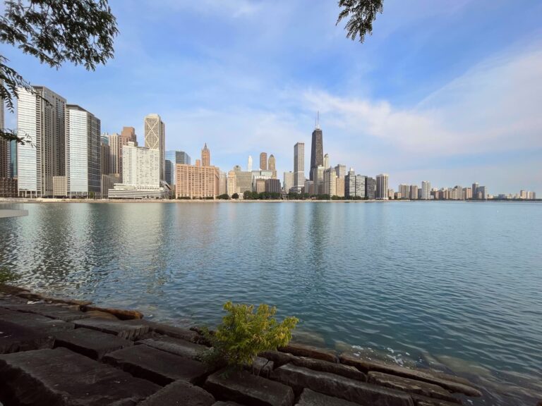 The Chicago skyline seen from the edge of a rocky waterfront, with calm blue waters and lush greenery. Tall modern buildings are stretched across the horizon under a clear blue sky.