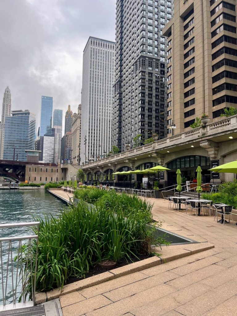 Chicago Riverwalk featuring sleek modern buildings and outdoor dining areas with green umbrellas beside the calm water. Lush plants line the walkway, creating a refreshing urban riverside setting.