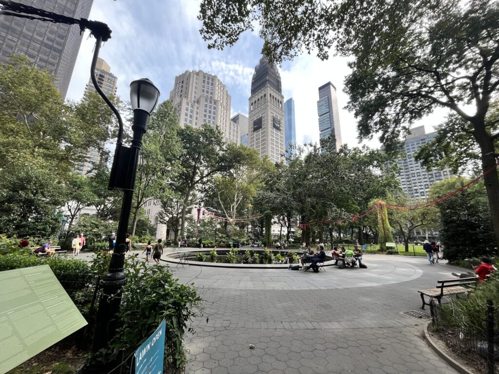 A scenic urban park with tall skyscrapers in the background surrounded by lush greenery. People are gathered around a circular fountain, enjoying the peaceful ambiance of the park.