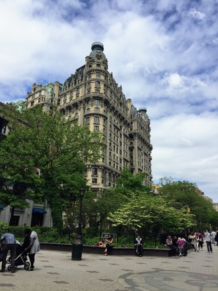 A stately, ornate stone building with green copper detailing at the top, surrounded by trees and bushes. People are relaxing on benches and walking through the pleasant, open urban space.