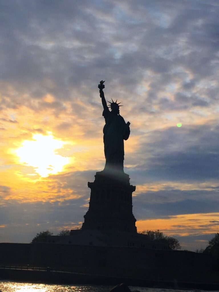 The Statue of Liberty silhouetted against a glowing orange and blue sunset. The iconic statue stands tall with its torch held high, symbolizing freedom and hope.
