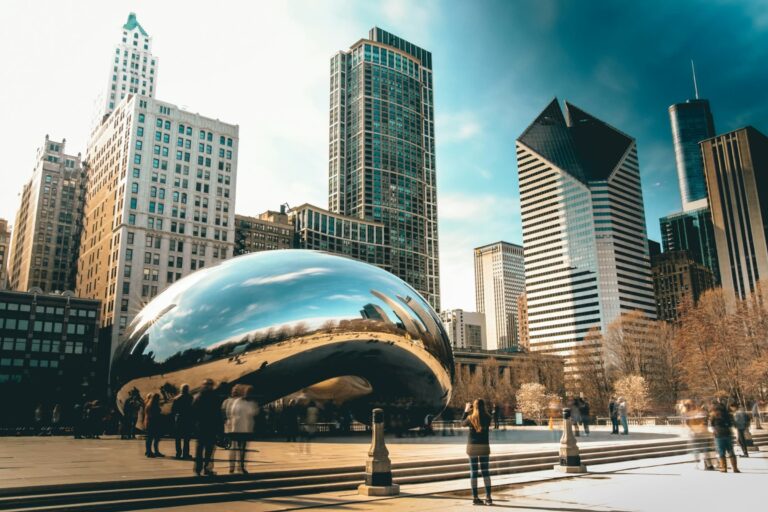 The Bean (Cloud Gate) sculpture in Chicago’s Millennium Park, reflecting the city’s skyscrapers and bright blue sky. Tourists gather around, taking photos of the shiny, curved surface.