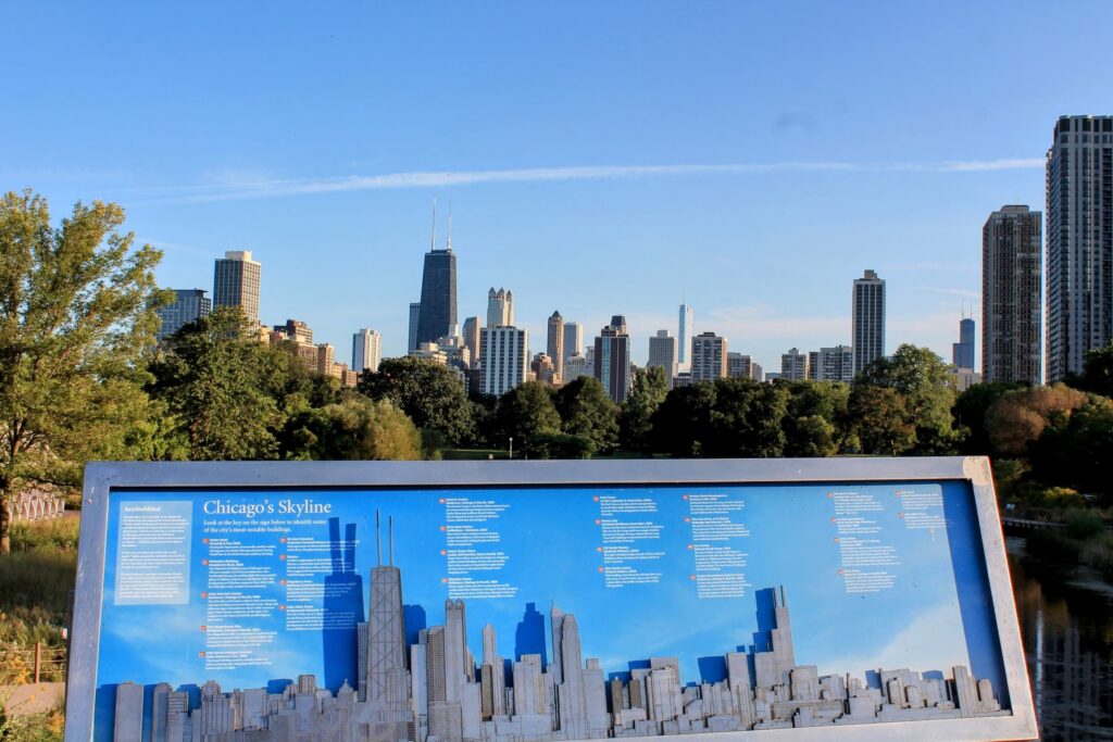 A Chicago skyline viewpoint featuring an informational sign that identifies notable buildings. The John Hancock Center and other skyscrapers rise above lush trees under a clear blue sky.