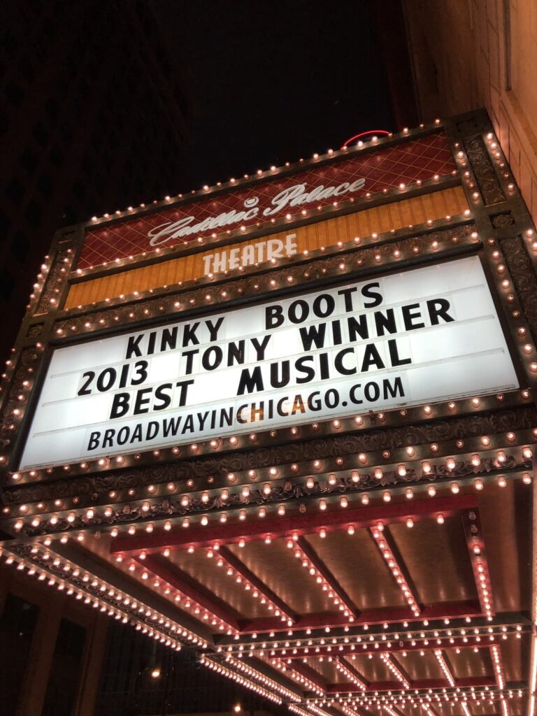 A glowing theater marquee at night reading "Kinky Boots - 2013 Tony Winner - Best Musical" at the Cadillac Palace Theatre in Chicago. The sign is illuminated by rows of bright, classic theater lights.