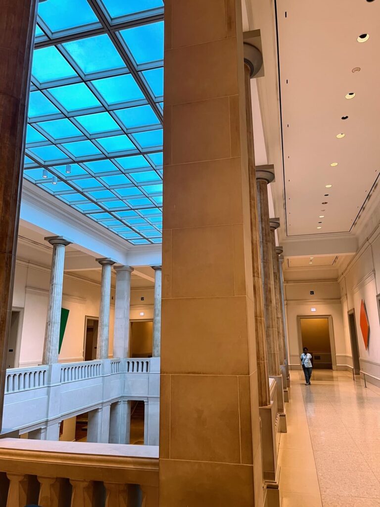 The grand interior of the Art Institute of Chicago, featuring high ceilings, classical columns, and a blue-tinted glass skylight. A lone visitor walks through the hallway, surrounded by elegant architecture.