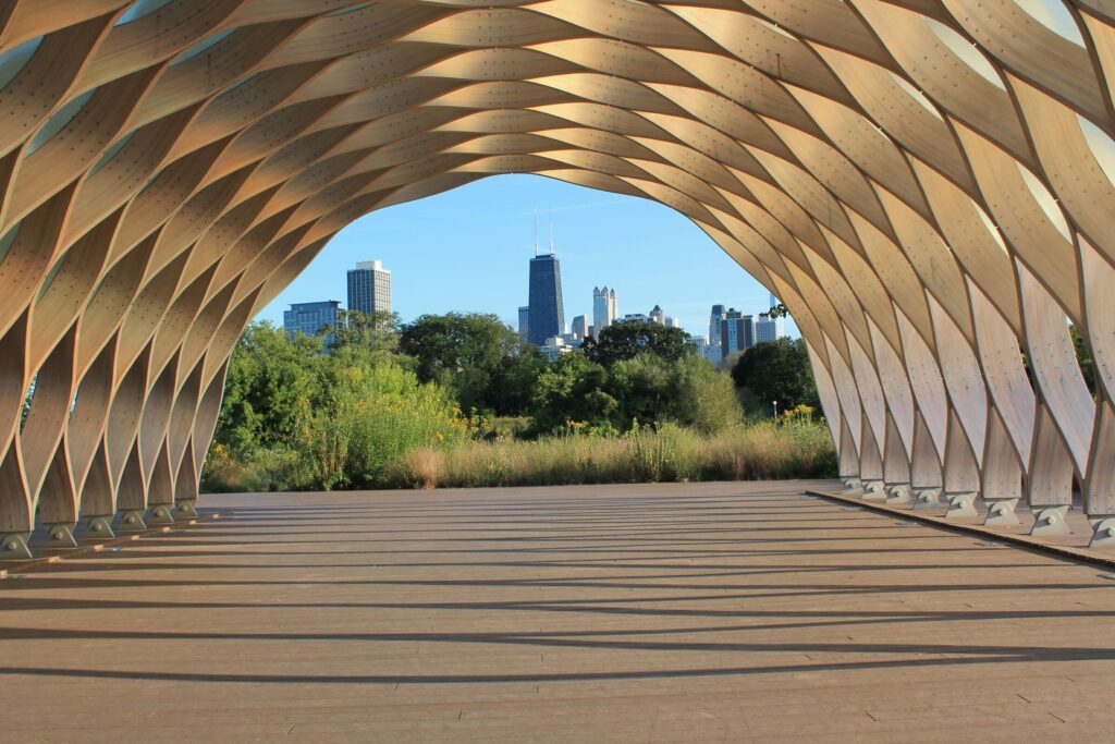 A stunning perspective from under the honeycomb-like wooden pavilion at Lincoln Park in Chicago. The arched structure frames a view of the city skyline, including the John Hancock Center, with lush greenery in the foreground.
