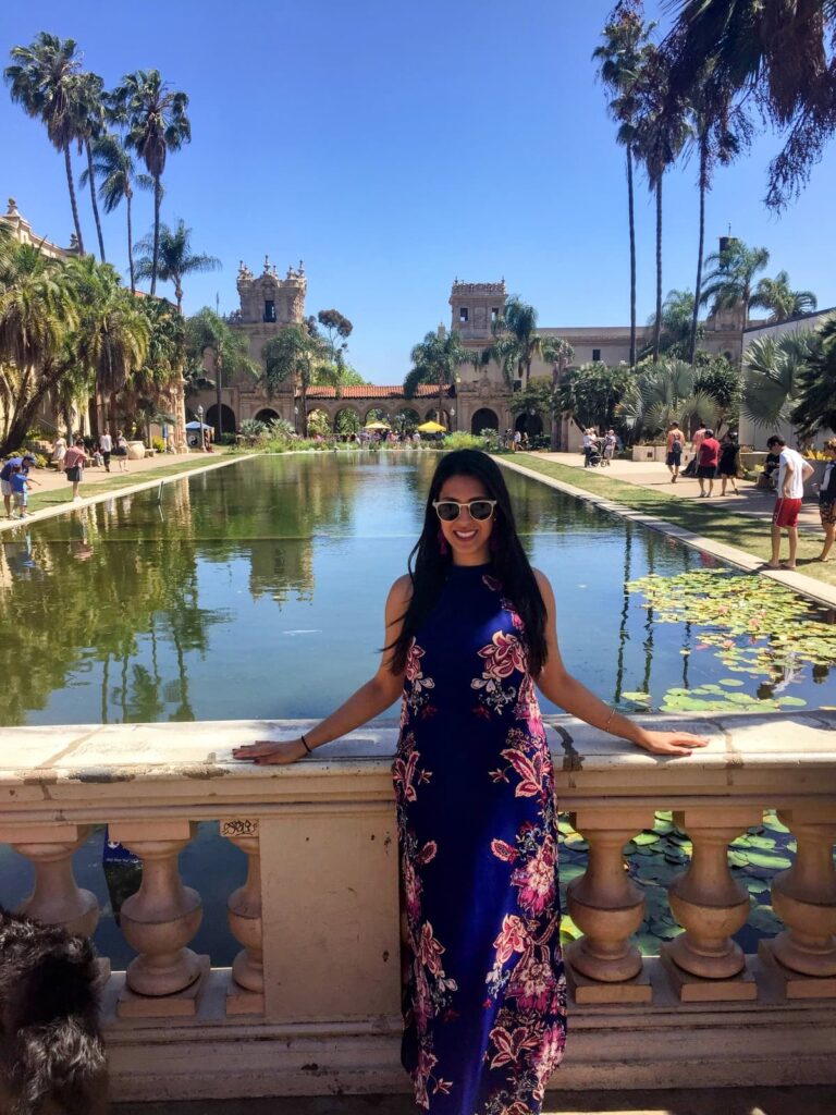 A woman in a long floral dress poses by a lily pond at Balboa Park, surrounded by Spanish-style buildings, palm trees, and clear reflections in the water.