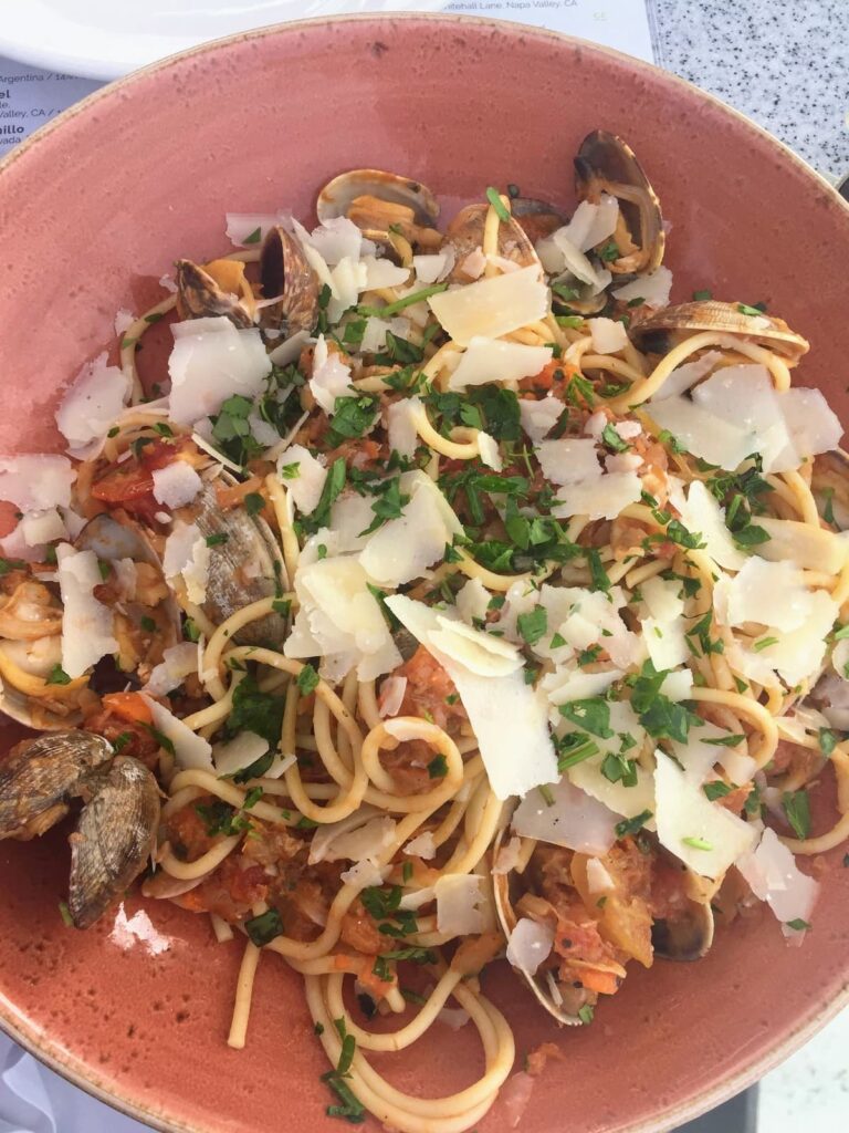 A seafood pasta dish in a terracotta bowl, topped with clams, tomato sauce, fresh parsley, and parmesan shavings.