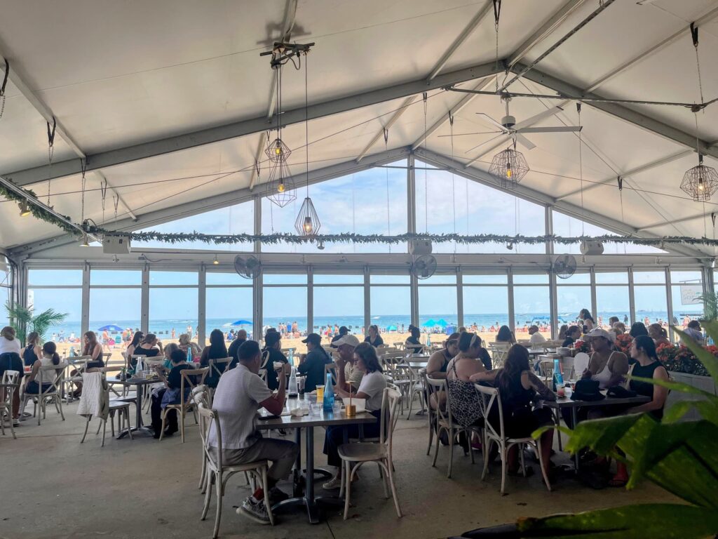 Indoor beachfront restaurant with people dining at tables, large windows showing beach umbrellas and a crowd enjoying the lakefront view.