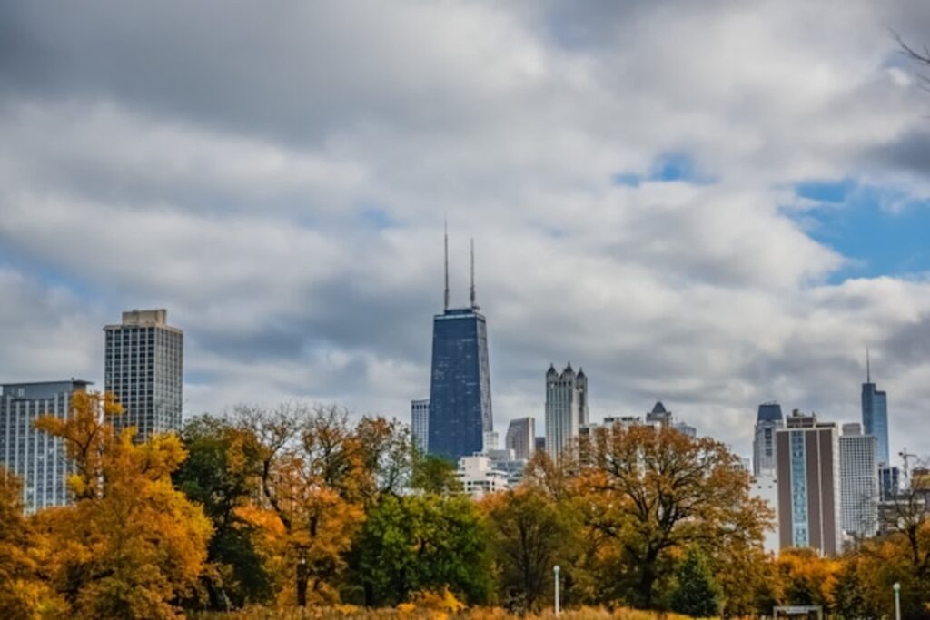 Chicago’s skyline peeks over a blanket of golden autumn trees, with the iconic John Hancock Center standing tall under a dramatic cloudy sky.