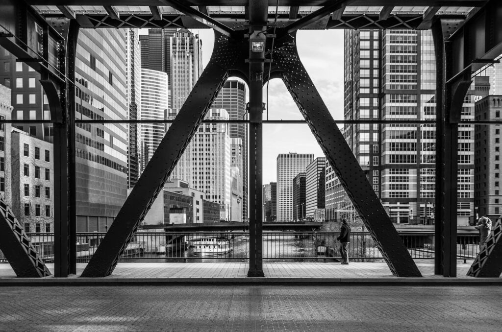 Black and white photo of a cityscape taken from a steel bridge, with large diagonal support beams framing a view of high-rise office buildings and skyscrapers. A river runs through the middle, with a docked tour boat below and two pedestrians walking along the bridge’s edge.