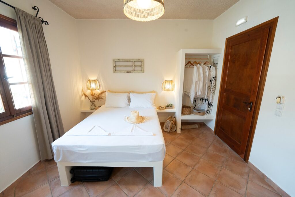 Bright, minimalist bedroom with a Mediterranean style, featuring a white double bed, two woven wall lamps, and an open wardrobe with neutral-toned clothing. The room has terracotta tile floors, light decor accents, and a window with soft beige curtains.