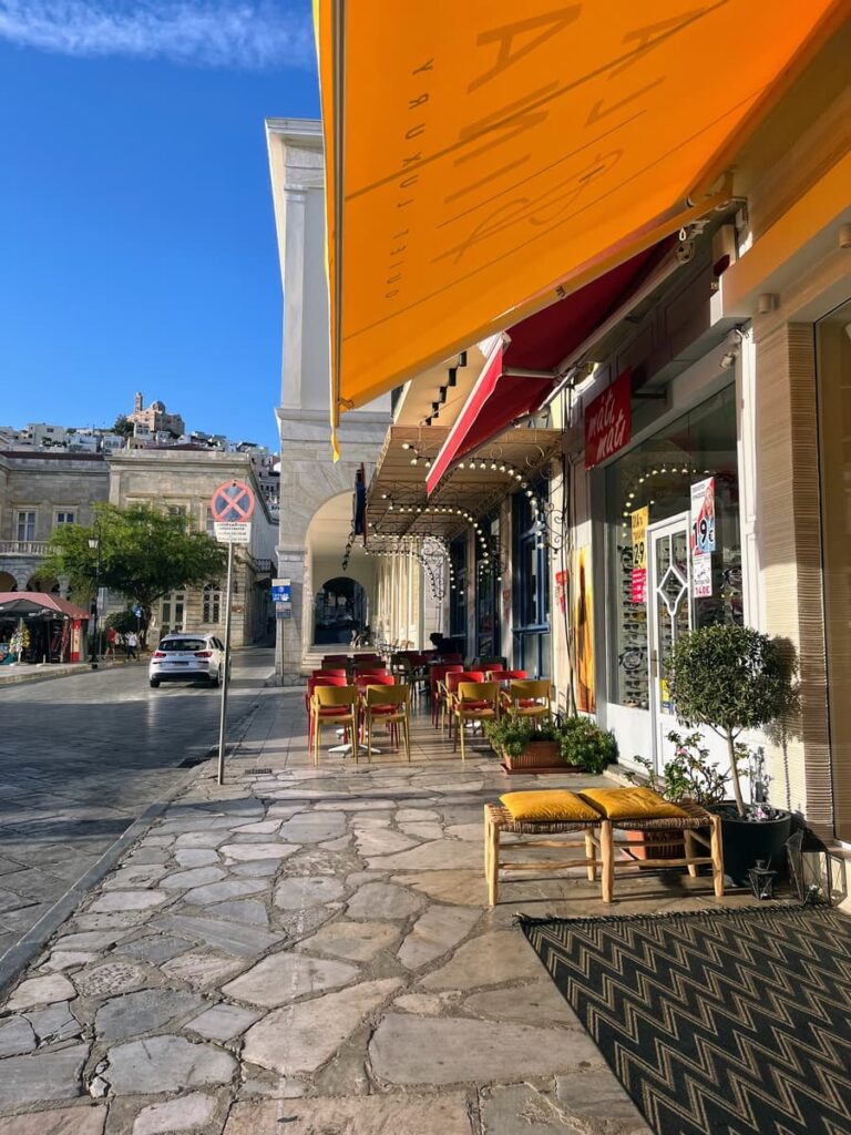 Main town - where to stay in Syros Greece Outdoor café seating along a stone-paved sidewalk, with bright yellow and red awnings providing shade to red and yellow chairs and small tables. In the distance, a church sits atop a hill, and the sky is a vivid blue with a thin cloud streak.