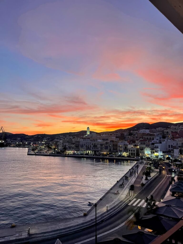 Colorful sunset over a harbor town, with soft hues of pink, orange, and purple reflecting on the calm water. The shoreline is lined with buildings and streetlights beginning to glow as the day transitions into night.