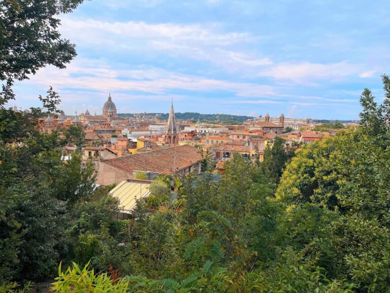 A wide, elevated view of Rome's rooftops and church domes framed by green trees, showcasing the city’s architectural beauty under a partly cloudy sky. post about digital nomad visa Italy process