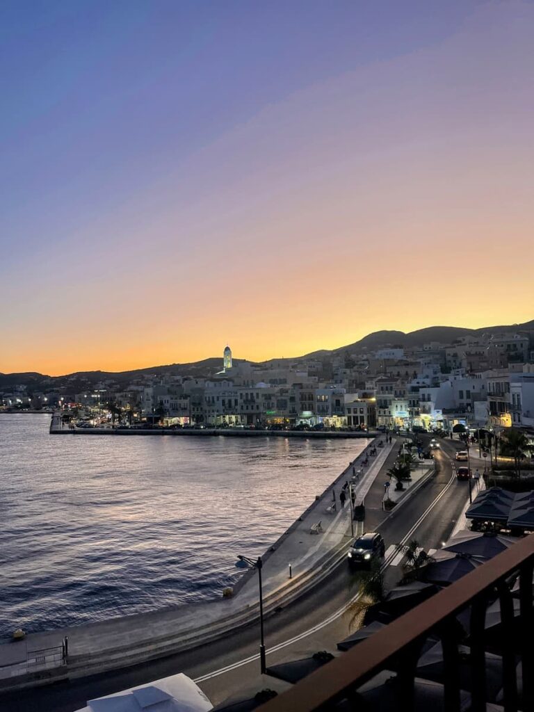 A coastal view of Syros harbor at twilight with golden and purple hues in the sky, city lights beginning to glow, and a curving road along the waterfront.