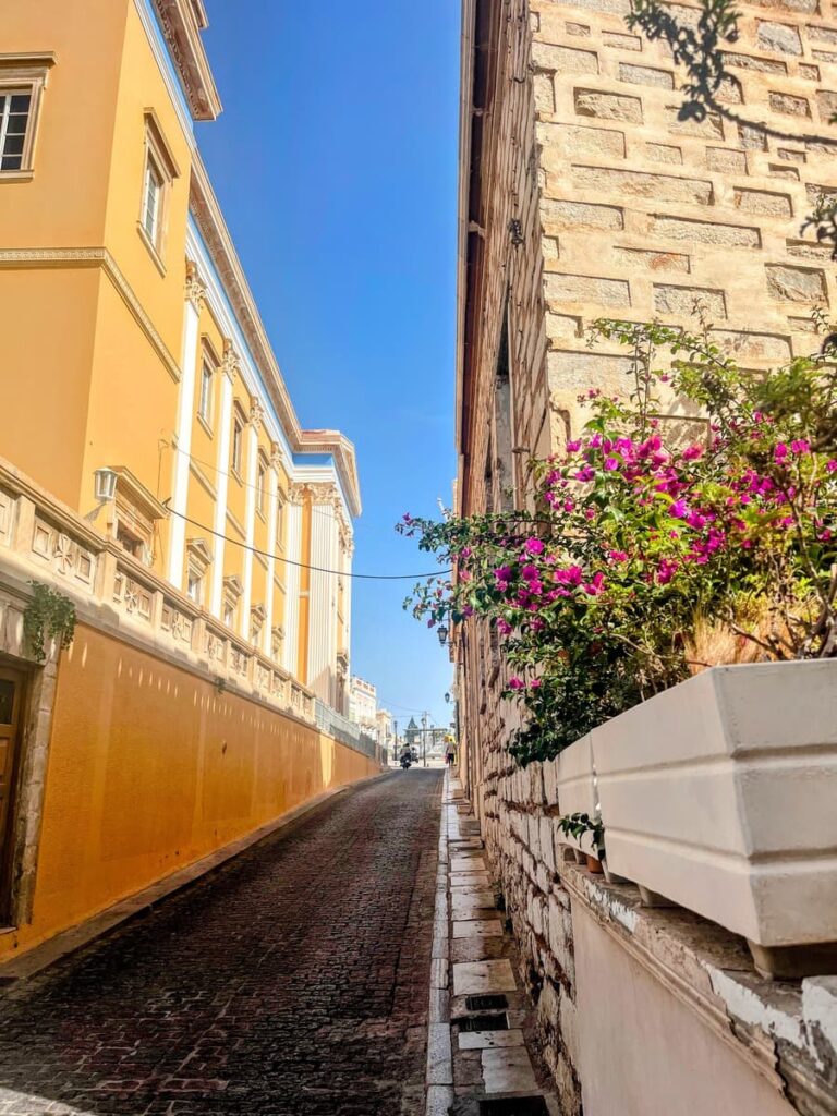 Narrow cobblestone street in bright sunlight with a yellow neoclassical building on the left and a stone building with blooming pink flowers in a planter on the right. The alley gently slopes uphill toward a blue sky.