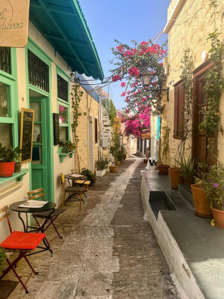 Charming alleyway with cobblestone pavement, lined with colorful buildings, potted plants, and small café tables. Bright pink bougainvillea arches over the narrow street under a clear blue sky.
