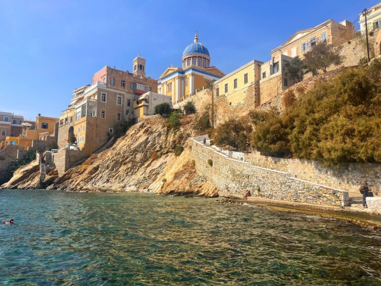 Seaside view of historic buildings on a rocky cliff, with a domed blue church as a focal point. The calm, clear water meets stone steps and a winding coastal path where a few people are walking or swimming. A great neighborhood for where to stay in Syros Greece
