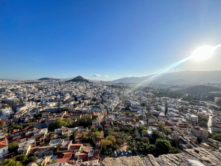 A panoramic view of Athens, Greece, taken from an elevated point at sunset, showing a dense spread of white buildings with Mount Lycabettus rising in the distance. The sun shines brightly in the upper right corner, casting a lens flare across the clear blue sky.