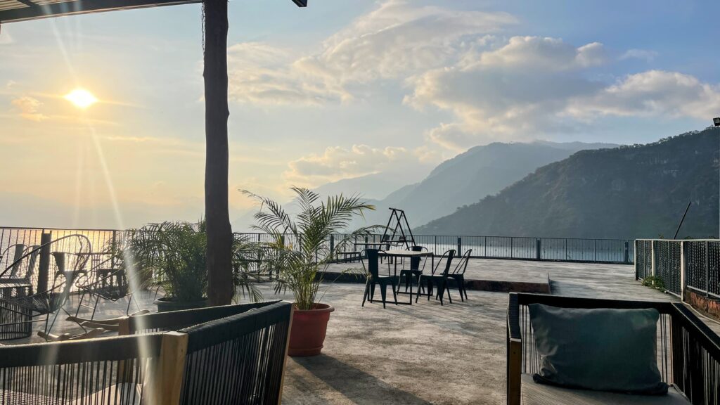 An open-air patio with modern seating overlooks a lake and misty mountains at sunset, with soft golden light casting long shadows across the concrete floor. A few potted plants and an empty swing set add to the peaceful atmosphere.