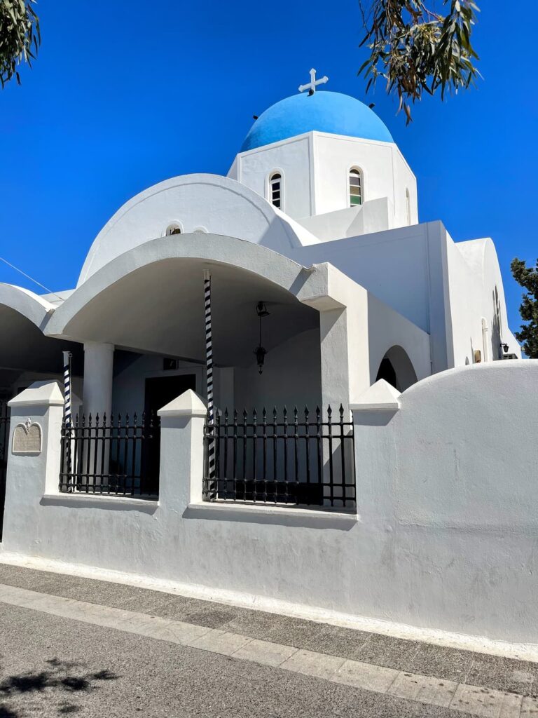 A white-walled Greek Orthodox church with a bright blue dome and cross stands out against a deep blue sky. The building features arched roofs and a black iron fence lining the street.