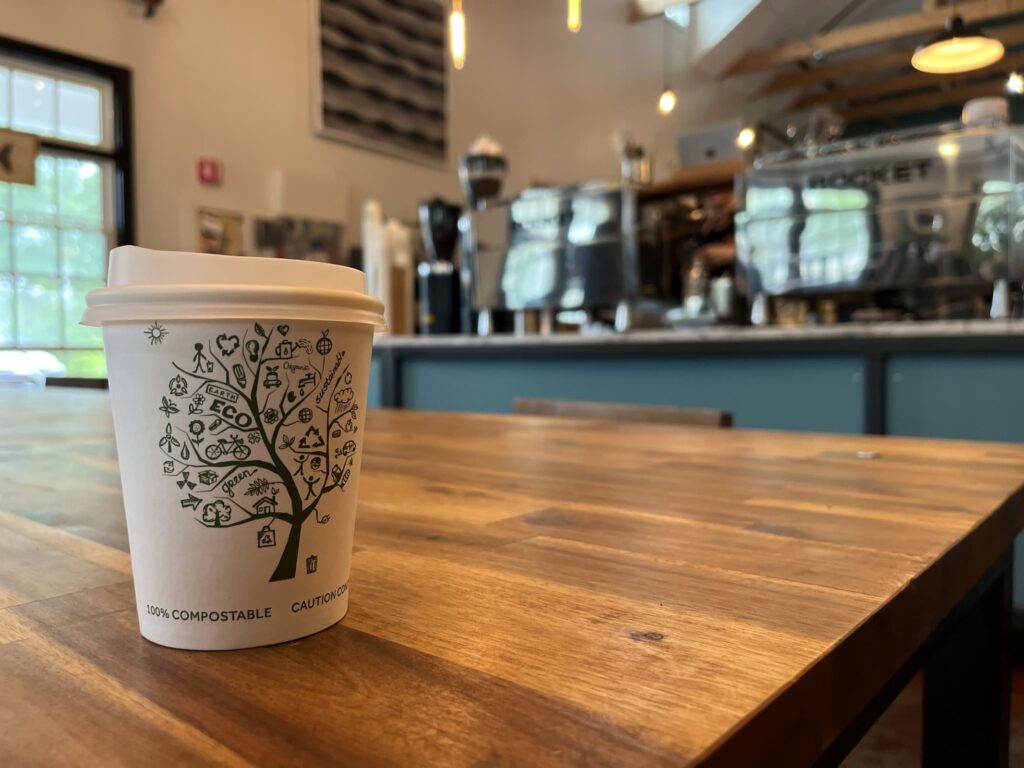 A compostable to-go coffee cup with eco-friendly illustrations sits on a wooden table in a cozy café, with espresso machines and baristas blurred in the background.
