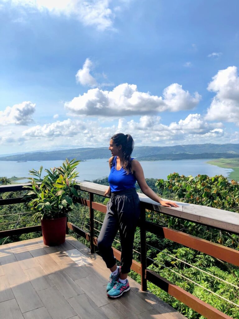 A woman in athletic wear stands on a scenic terrace overlooking a lush green valley and a large lake under a bright, partly cloudy sky. She leans on a wooden railing next to a potted plant, smiling toward the view.
