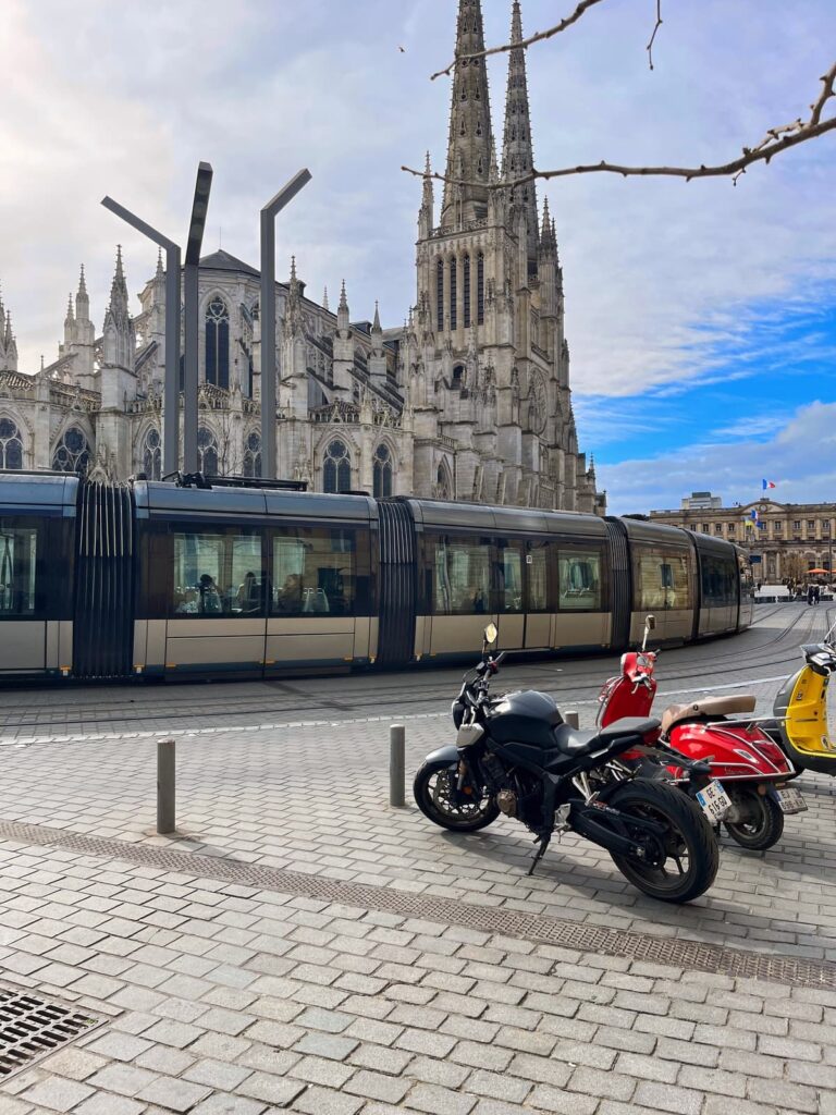 A modern tram passes in front of a gothic-style cathedral with twin spires in Bordeaux, France. Three parked scooters and a motorcycle sit in the foreground on cobblestone streets.
