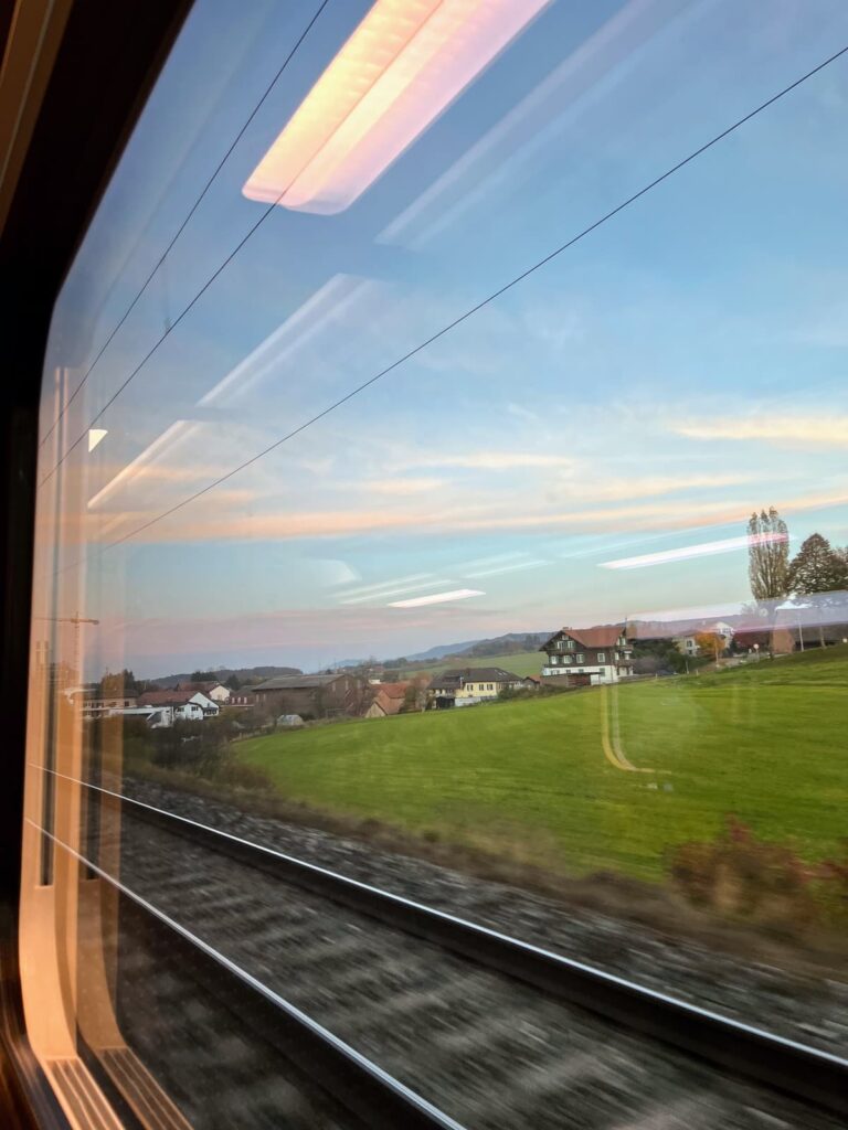 A view from inside a train window shows railway tracks and a green countryside with homes and hills in the distance. Reflections of overhead lights are visible on the glass.