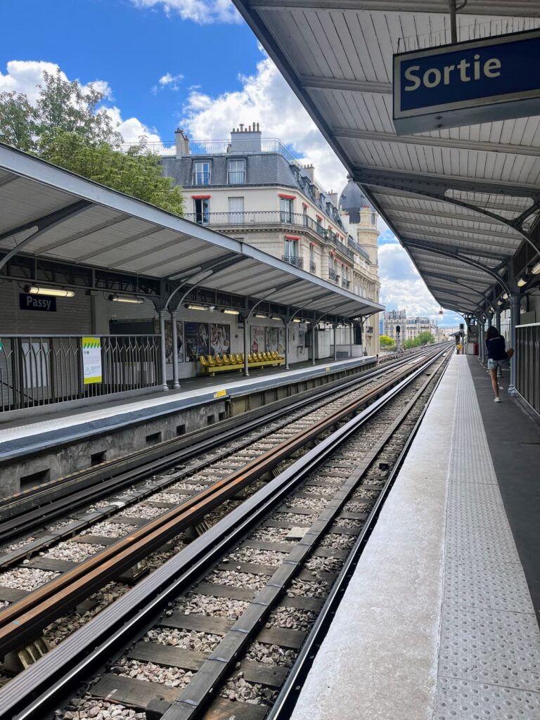 A quiet Paris Metro station platform with empty tracks and yellow seats sits under a cloudy sky. The station sign reads “Passy,” and an exit sign marked “Sortie” hangs from the roof.