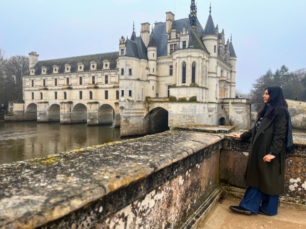 Large castle over a river, woman stands at a nearby lookout point wearing a large coat, scarf and boots in winter in the Loire Valley
