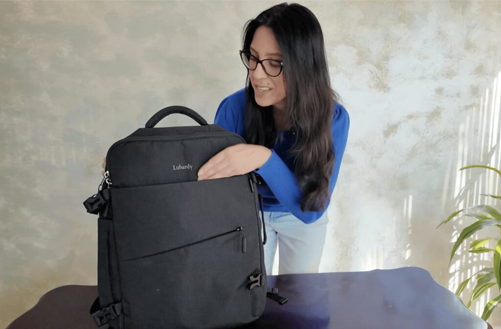 A woman in a blue sweater and glasses smiles while showing the front pocket of a black Lubardy travel backpack on a table. The scene is warmly lit with a neutral wall background.
