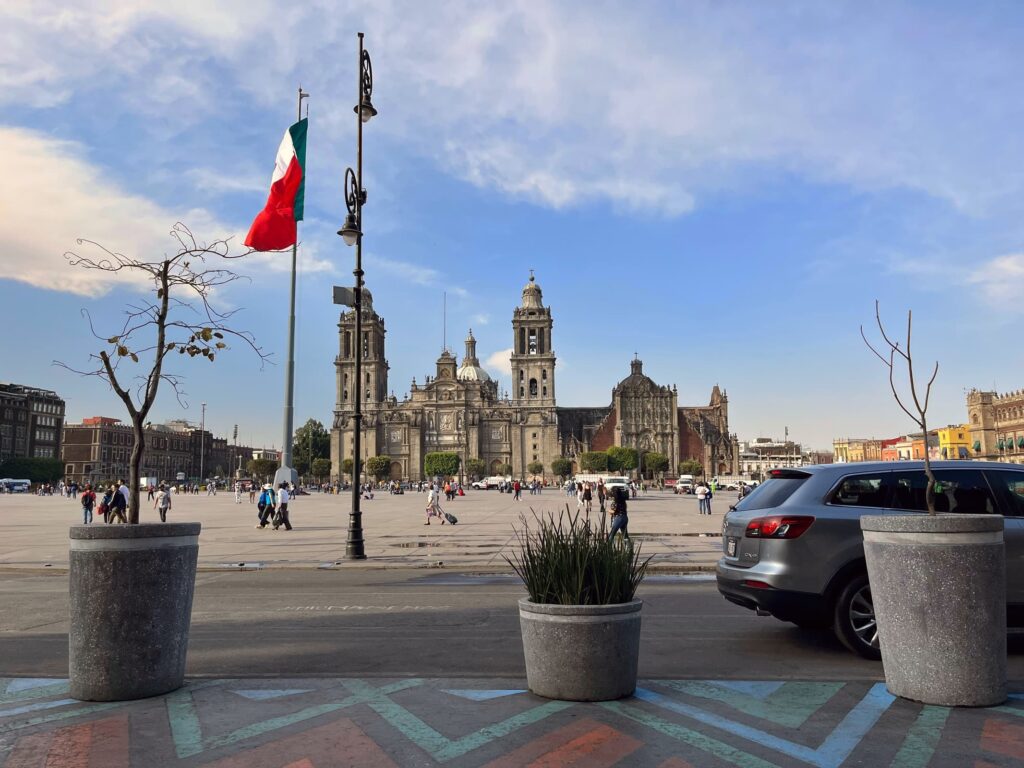 View of the historic Zócalo square in Mexico City with the Metropolitan Cathedral in the background, a large Mexican flag waving in the center, and people walking across the wide open plaza.