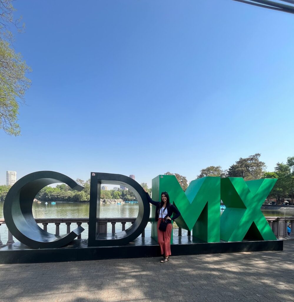 A woman poses next to the large black and green CDMX sign near a lake in Chapultepec Park, with trees, paddle boats, and city buildings visible in the distance.