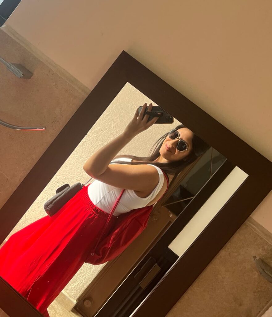 A woman takes a mirror selfie indoors, wearing sunglasses, a white top, and a flowing red skirt with a red backpack, standing in front of a wooden-framed mirror.