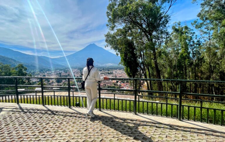 Woman dressed in white stands at a scenic overlook, gazing at a distant volcano framed by sun rays and blue skies, with the city of Antigua, Guatemala sprawling below. Tall trees line the viewpoint, adding to the serene and elevated perspective.
