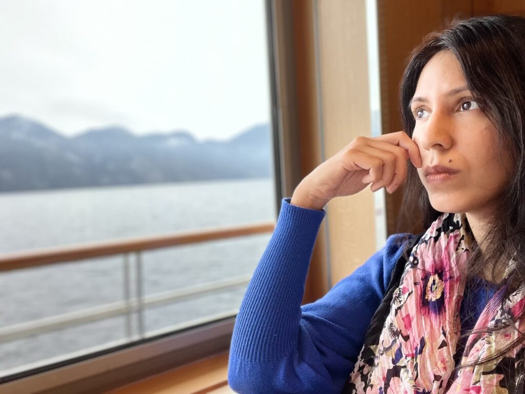 Woman wearing a blue sweater and floral scarf gazes out the window of a boat, with a calm lake and misty mountains in the background.