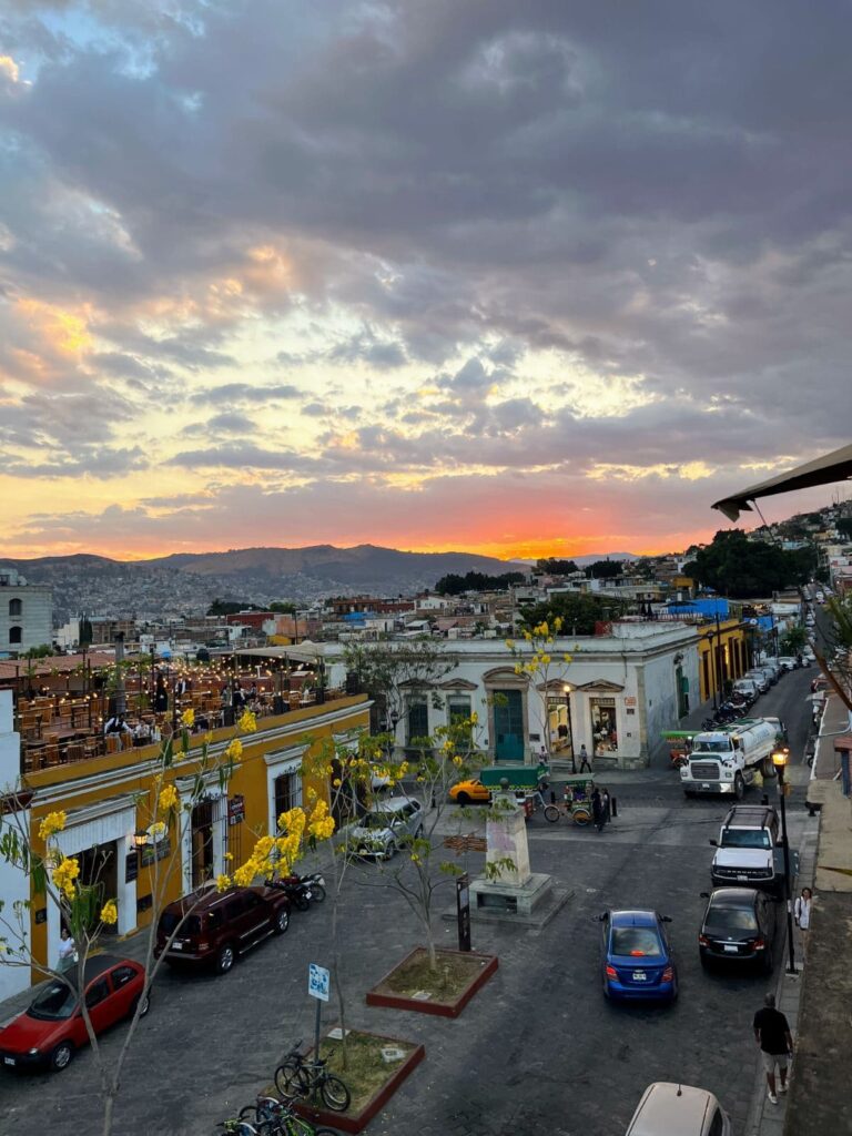 Colorful sunset over the colonial buildings and vibrant street life of Oaxaca, Mexico, with yellow flowering trees and a rooftop restaurant visible.