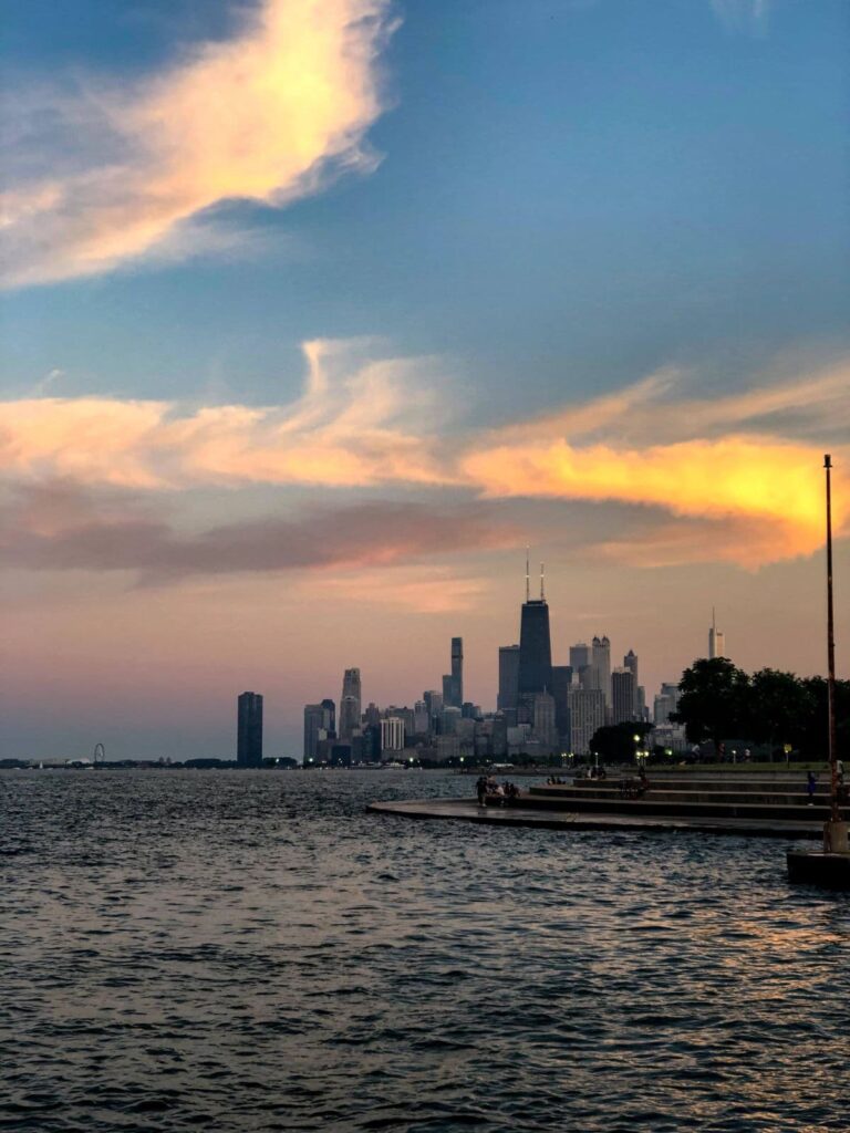 View of the Chicago skyline at sunset from the edge of Lake Michigan, with soft pastel clouds above and the John Hancock Center visible in the distance.