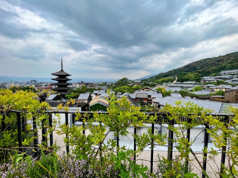 Scenic view over Kyoto’s traditional tiled rooftops with a five-story pagoda rising in the distance, framed by lush green hills and dramatic cloudy skies.