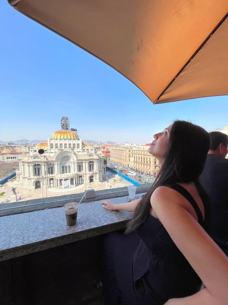 A woman seated at a rooftop café gazes out toward the Palacio de Bellas Artes in Mexico City, with an iced drink on the counter and a wide cityscape in view.